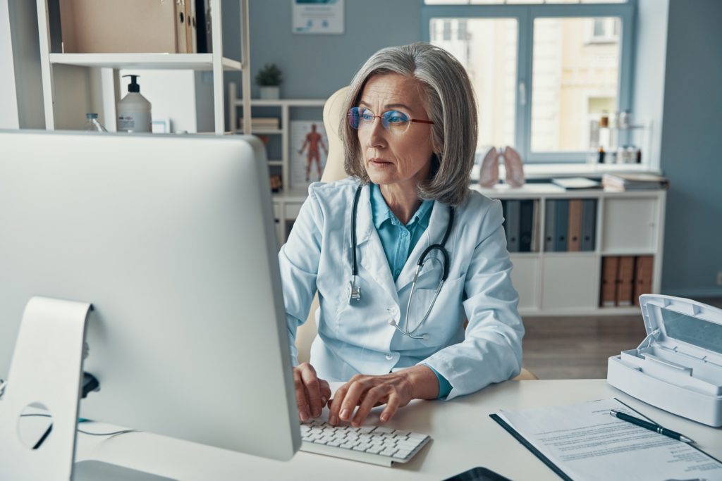 Serious mature female doctor in white lab coat working using computer while sitting in her office